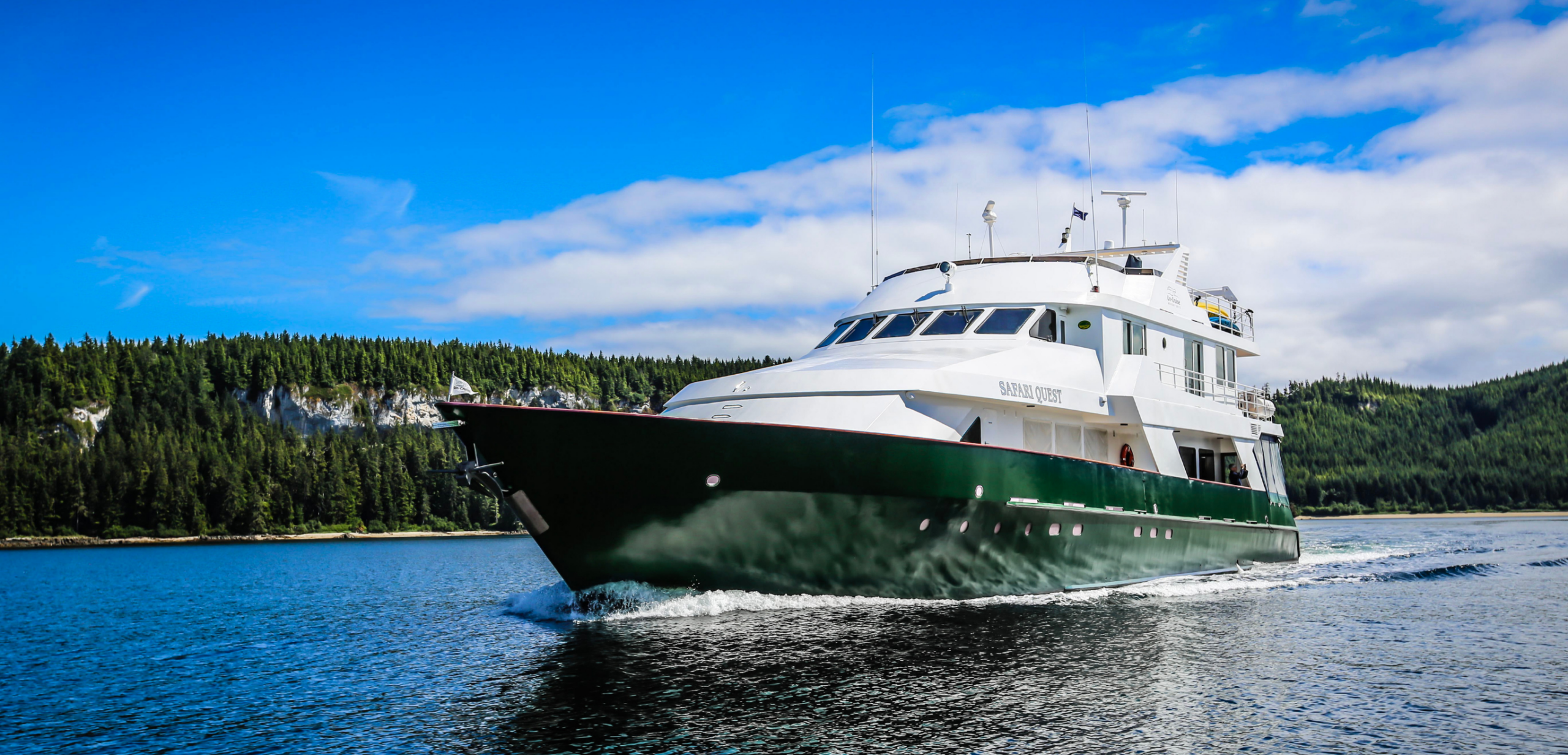 Safari Quest Expedition Yacht in Alaska A luxurious small expedition yacht named Safari Quest cruises through pristine Alaskan waters, surrounded by dense evergreen forests and rocky coastline. The vessel features distinctive white upper decks and dark green hull, designed for intimate wildlife and glacier exploration in remote northern regions.
