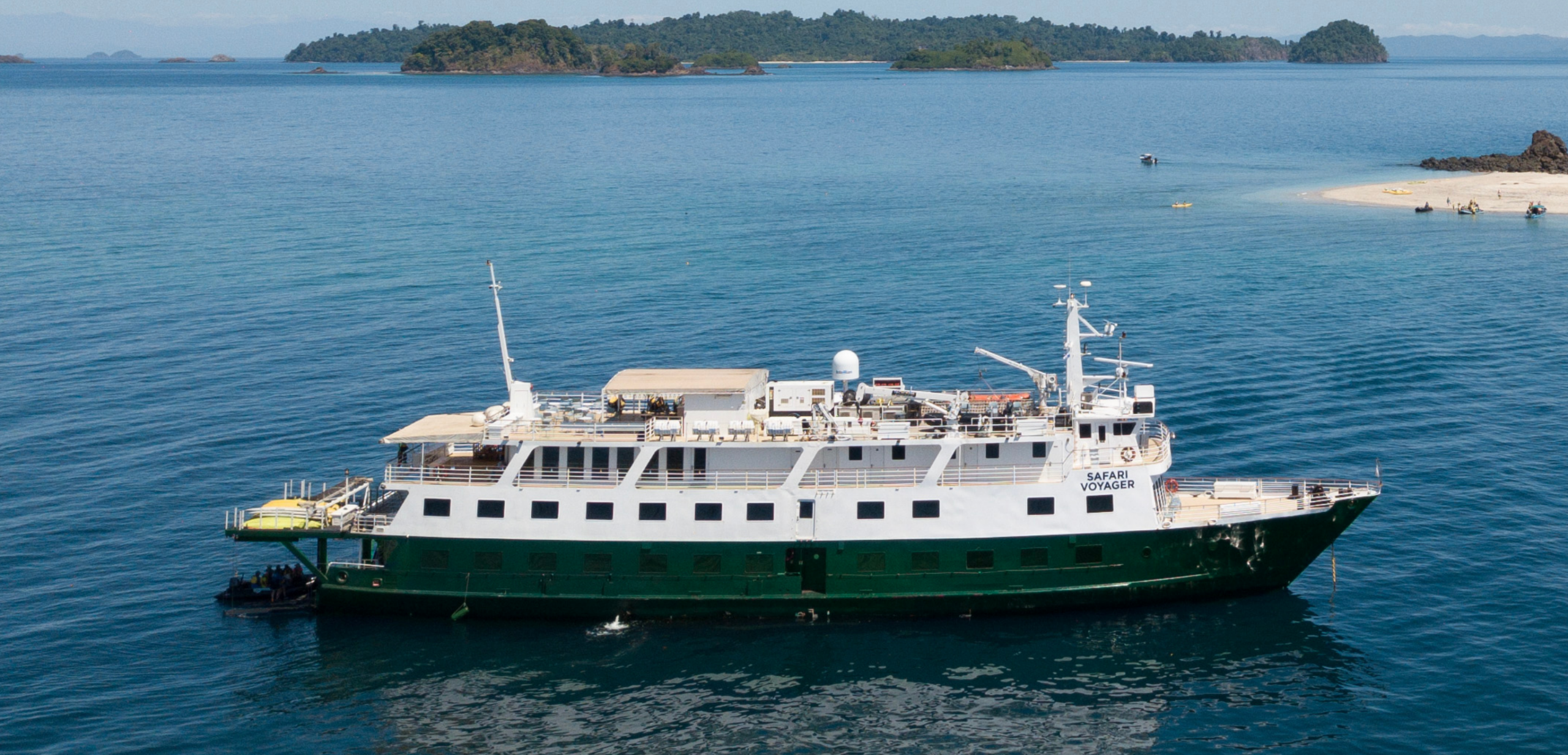 Safari Voyager Anchored in Tropical Waters The Safari Voyager, a small luxury expedition cruise ship, is anchored in clear turquoise waters off a pristine tropical island with lush forested terrain and sandy beaches. The white and green vessel is positioned near a shallow beach area with small boats and passengers visible, showcasing the intimate, destination-focused nature of expedition cruising.