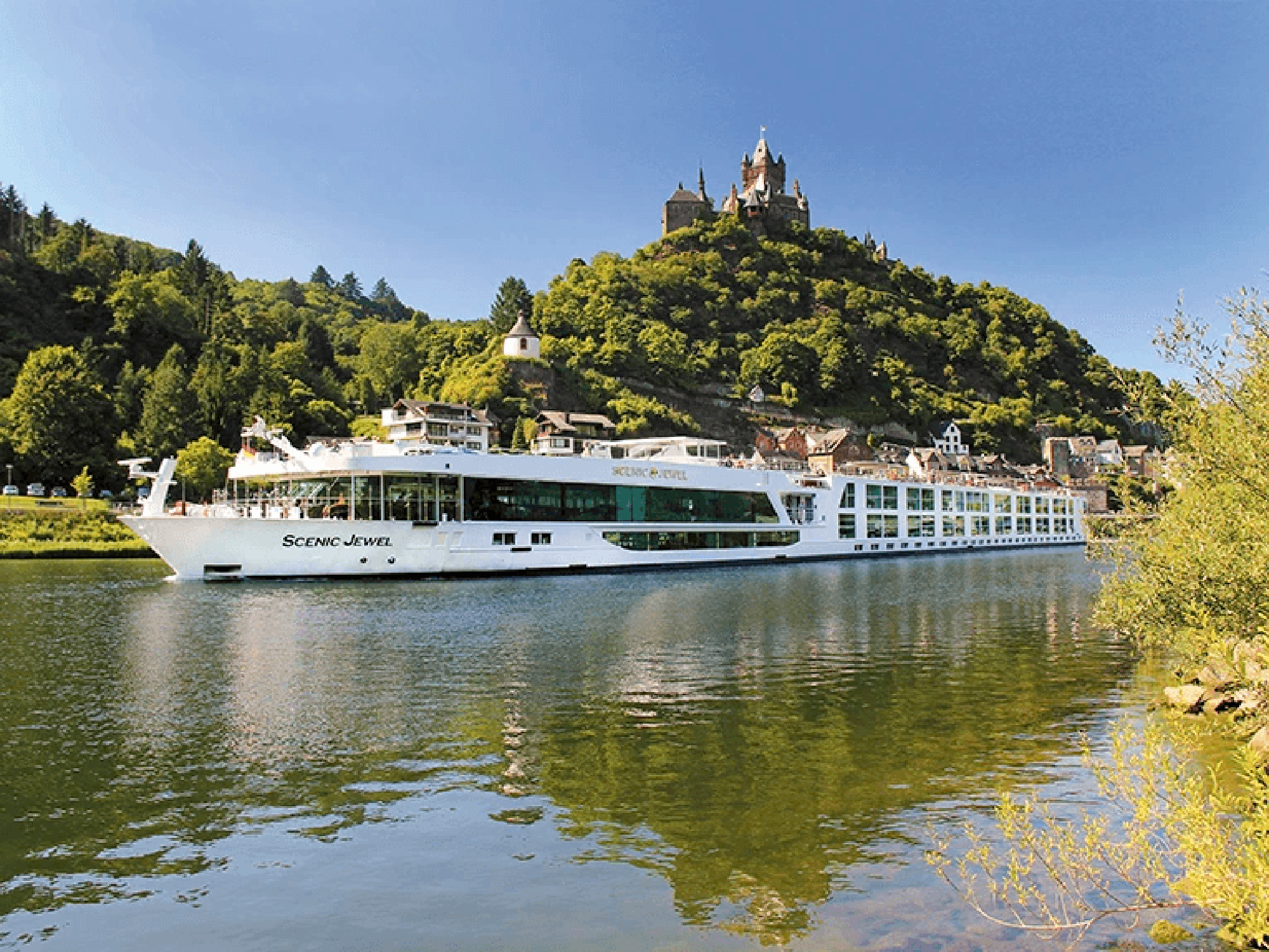 Scenic Jewel River Cruise Near Neuschwanstein Castle The Scenic Jewel river cruise ship navigates a picturesque European river with the iconic Neuschwanstein Castle perched dramatically on a forested hilltop in the background. The modern riverboat features extensive glass windows and contemporary design, contrasting beautifully with the medieval castle and charming riverside village architecture.