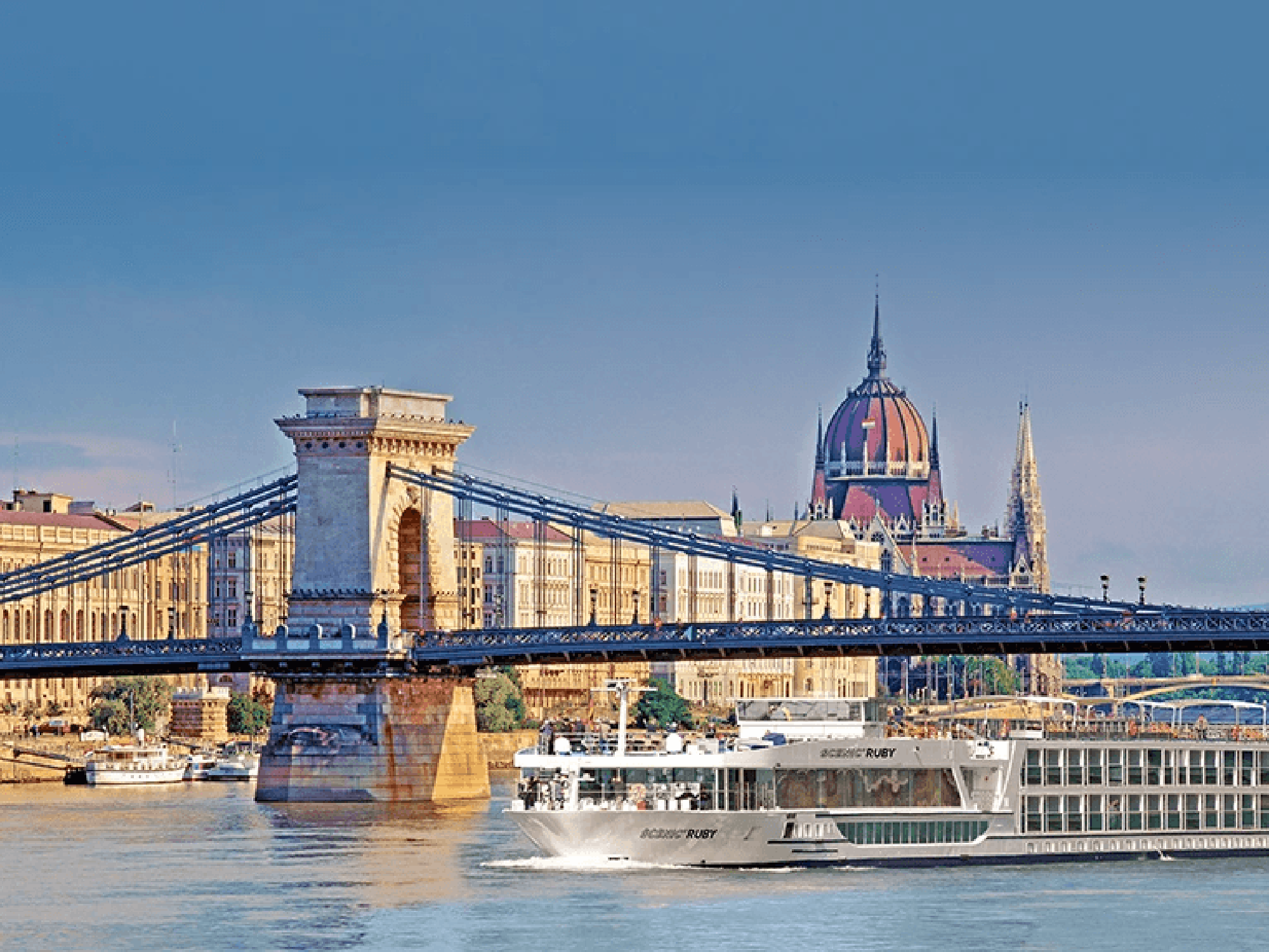 Scenic Ruby River Cruise Budapest A river cruise ship passes beneath the iconic Chain Bridge in Budapest, Hungary, with the stunning Hungarian Parliament Building and its distinctive red dome visible in the background. The image captures the golden-hour sunlight reflecting off the Danube River and the historic architecture of Budapest's skyline.