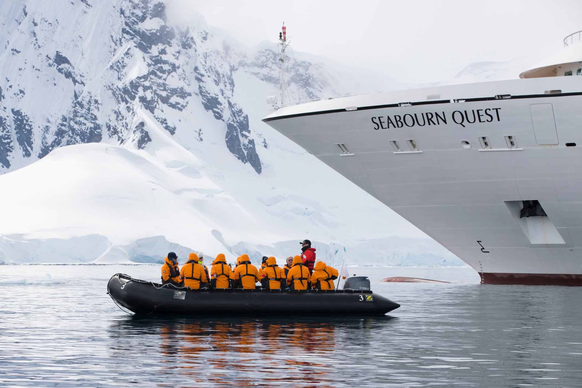 Passengers in bright orange life jackets explore Antarctic waters aboard a black Zodiac inflatable boat, with the luxurious Seabourn Quest cruise ship visible in the background against towering snow-covered mountains and glaciers. This image captures an adventurous polar expedition excursion in one of the world's most remote and pristine regions.