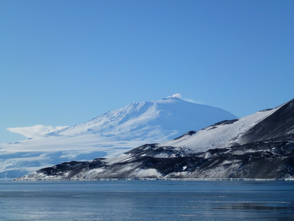 A majestic snow-covered volcanic mountain rises dramatically from an ice-laden Arctic shoreline, with pristine white peaks contrasting against a brilliant blue sky and dark volcanic rock formations in the foreground. The scene captures the raw, remote beauty of a polar landscape with frozen coastal waters and glaciated terrain.