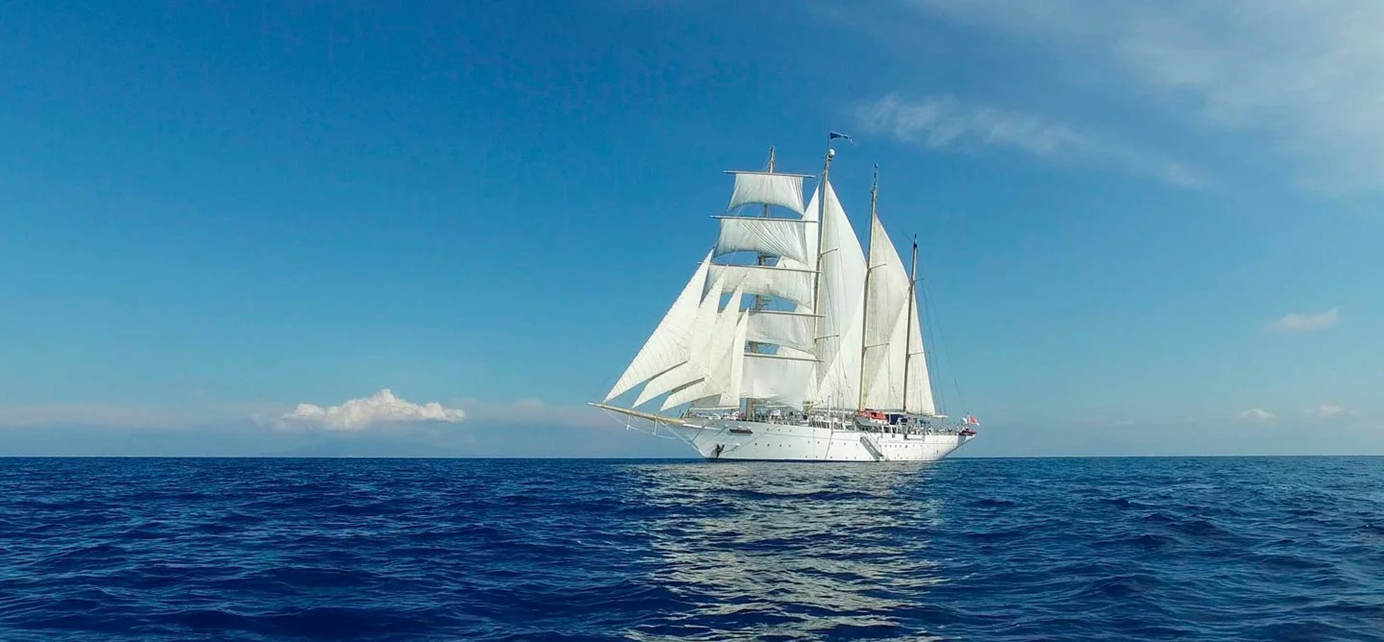 A magnificent white-hulled tall ship with full square-rigged sails deployed, sailing across calm blue ocean waters under a clear sky with scattered clouds. The vessel appears to be a modern sailing cruise ship, featuring multiple masts and traditional sailing rigging combined with contemporary maritime construction.