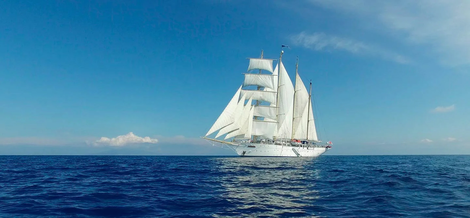 A stunning white-hulled tall ship with full square-rigged sails deployed sails across calm blue waters under a clear sky. The classic sailing vessel features multiple masts with billowing white canvas and a modern white superstructure, exemplifying traditional maritime elegance combined with contemporary cruise ship amenities.