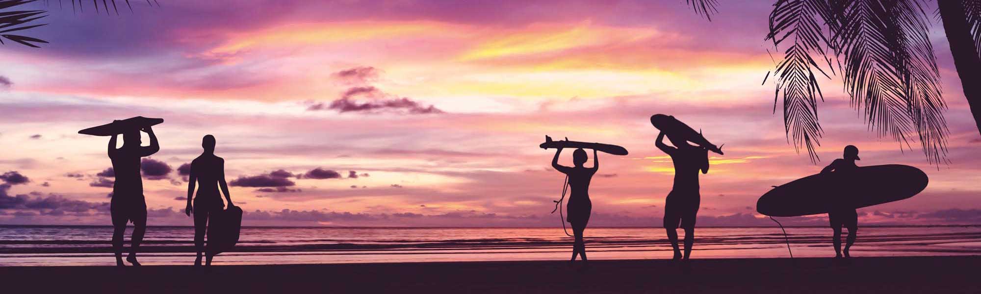 A striking silhouette composition of multiple surfers with their boards gathered on a tropical beach at golden hour, with vibrant pink and purple sunset skies reflected across calm waters and palm fronds framing the scene.