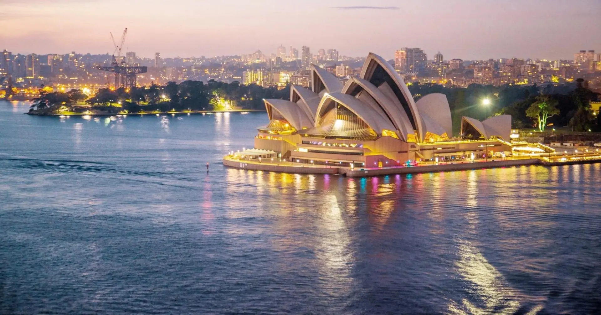 The iconic Sydney Opera House is brilliantly illuminated against the twilight sky, with its distinctive white shell-shaped structures reflecting in the calm waters of Sydney Harbour. The cityscape of Sydney glows in the background, creating a stunning evening portrait of this world-famous landmark and popular cruise destination.
