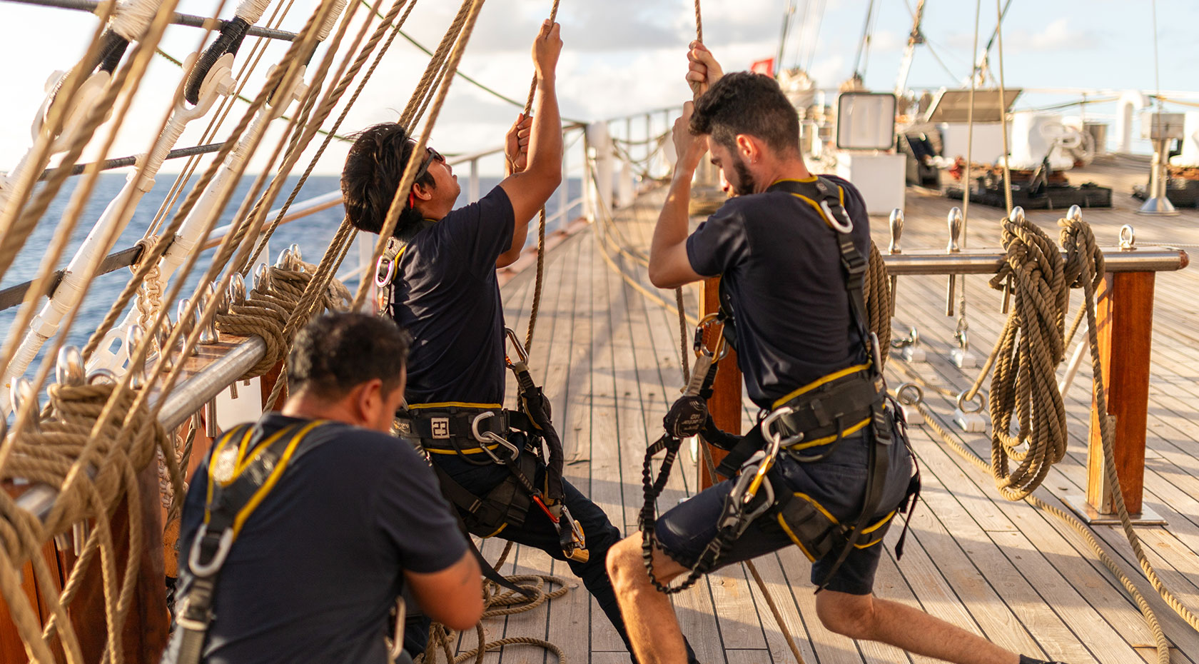 Tall Ship Crew Training Exercise at Sea Crew members in safety harnesses work together on the rigging and deck of a traditional tall ship, demonstrating coordinated maritime training and seamanship skills. The image captures the collaborative nature of sailing operations with multiple sailors managing ropes and rigging systems under blue sky conditions.