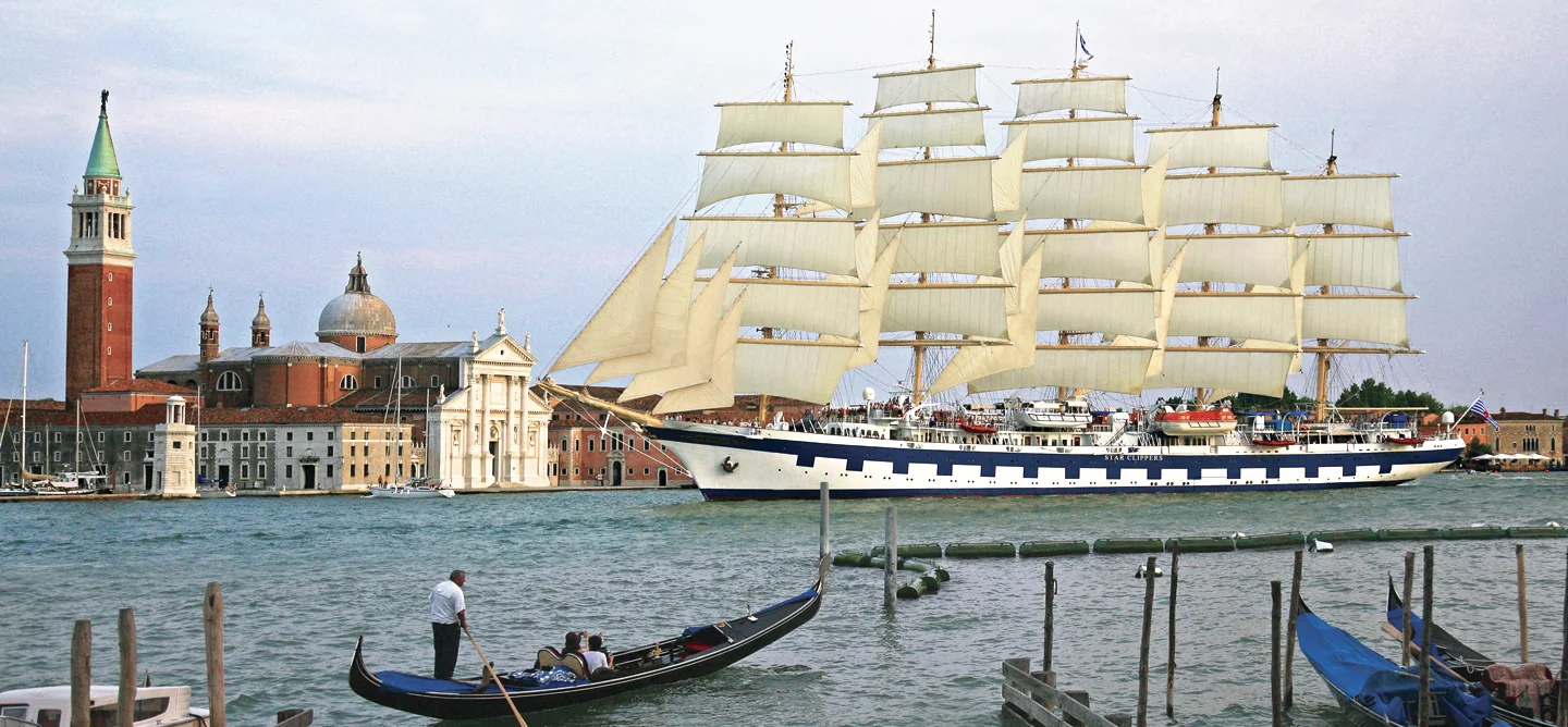 A magnificent full-rigged sailing vessel with billowing cream-colored sails navigates Venice's iconic Grand Canal, passing the historic San Giorgio Maggiore church and campanile. Traditional gondolas with mooring poles dot the foreground, showcasing the contrast between classic maritime heritage and Venetian waterfront architecture.