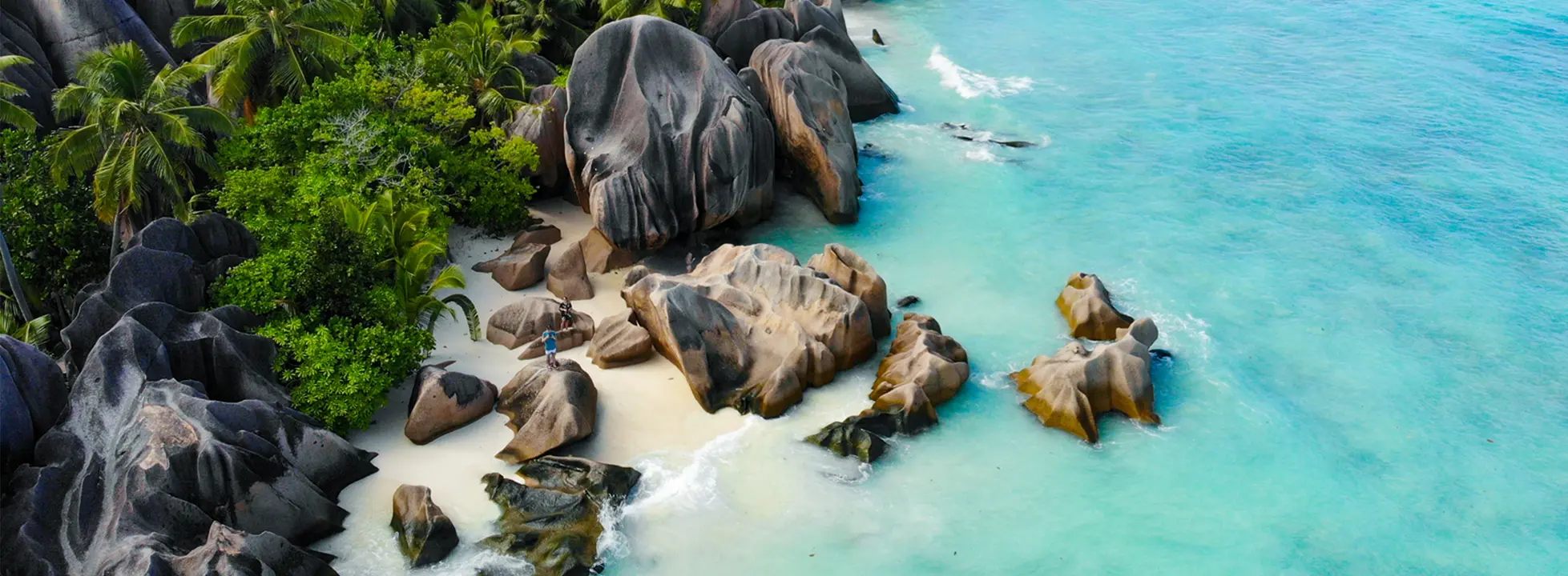 A stunning tropical beach cove featuring distinctive large granite boulders scattered along pristine white sand, bordered by turquoise waters and lush tropical vegetation. The dramatic rock formations create a picturesque natural landscape ideal for beach exploration and water activities.