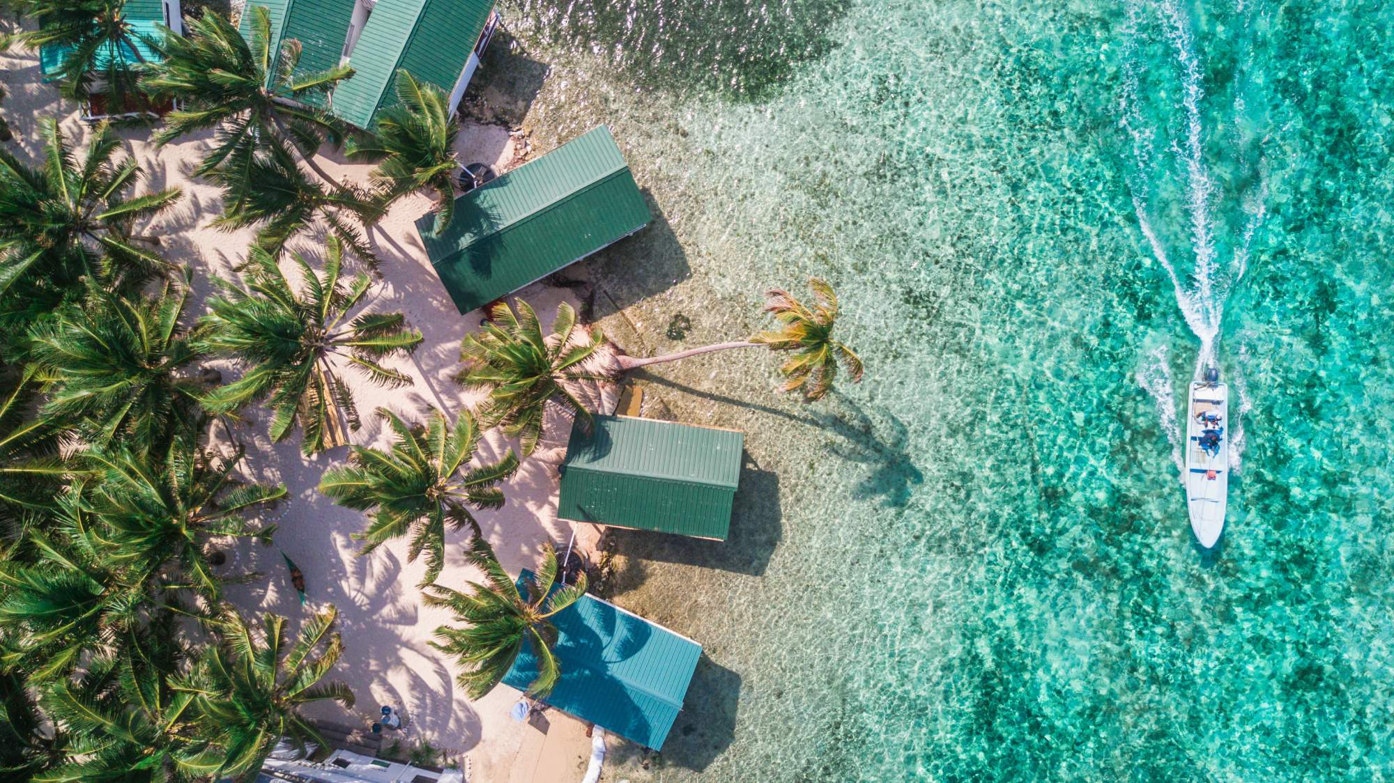 An aerial overhead view of a pristine tropical beach with turquoise waters, white sand, and several traditional thatched-roof bungalows with green metal roofing nestled among coconut palm trees. A small white fishing or tour boat is visible in the shallow turquoise waters offshore.