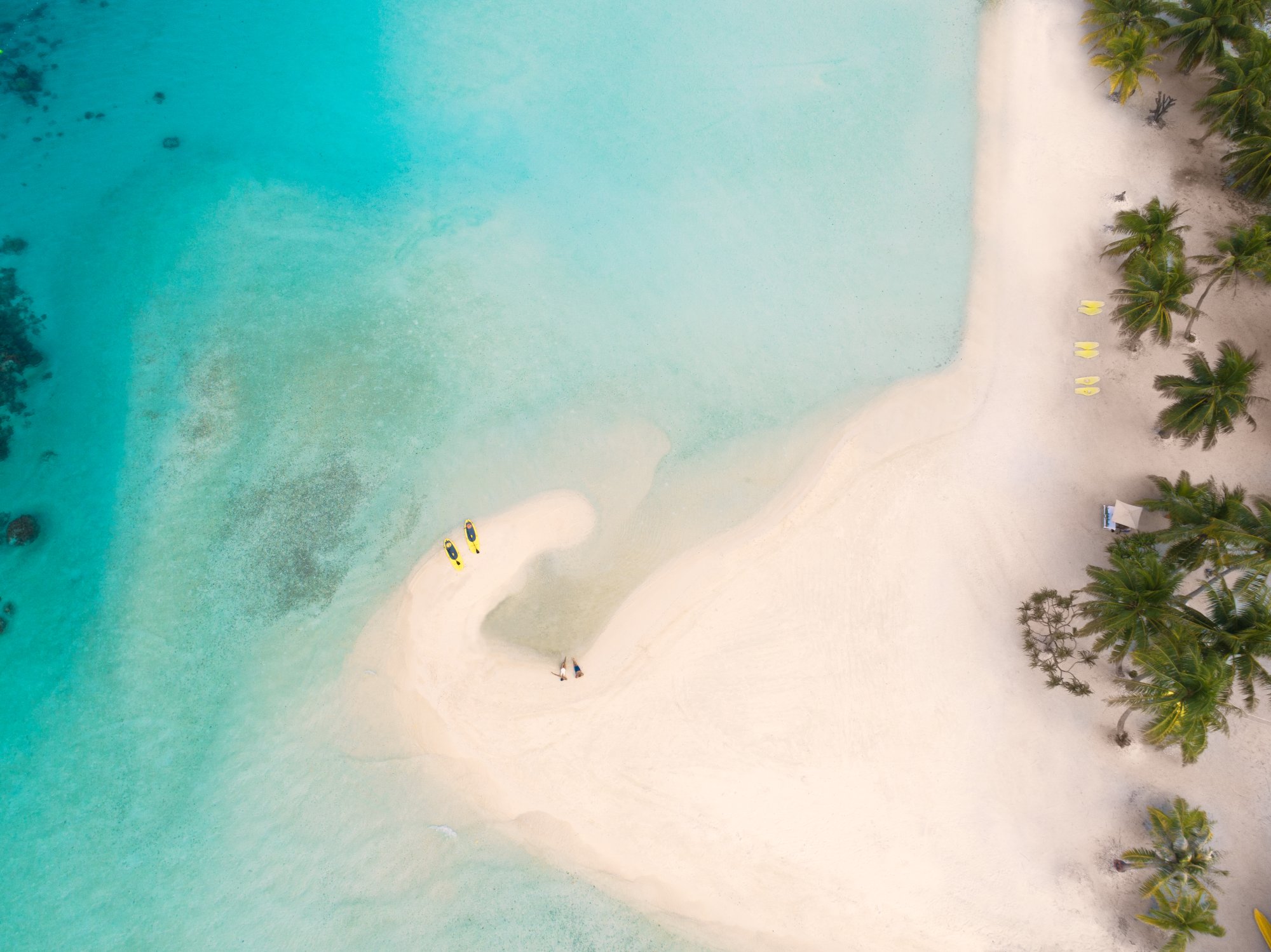 An aerial view of a pristine white sand beach in crystal-clear turquoise waters, featuring beachgoers, yellow kayaks, and palm trees along the shoreline. The shallow lagoon displays vibrant aquamarine hues characteristic of French Polynesian atolls.
