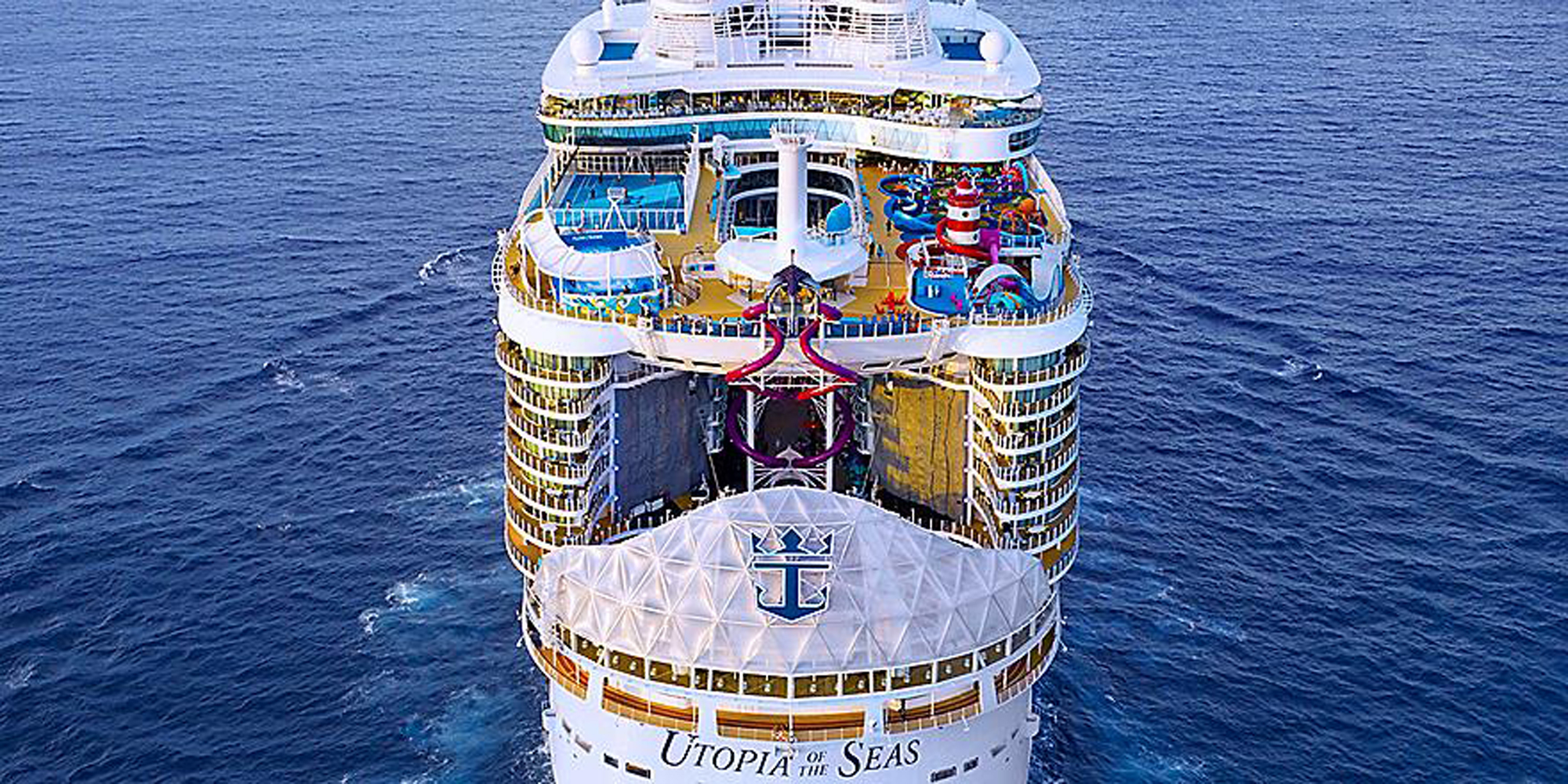 An aerial bow-on view of the Utopia of the Seas cruise ship at sea, displaying its distinctive white hull, multiple colorful deck levels with railings, and iconic branding visible on the ship's name plaque. The vessel showcases multiple entertainment decks with visible pools, recreational areas, and holiday decorations, surrounded by deep blue ocean water.