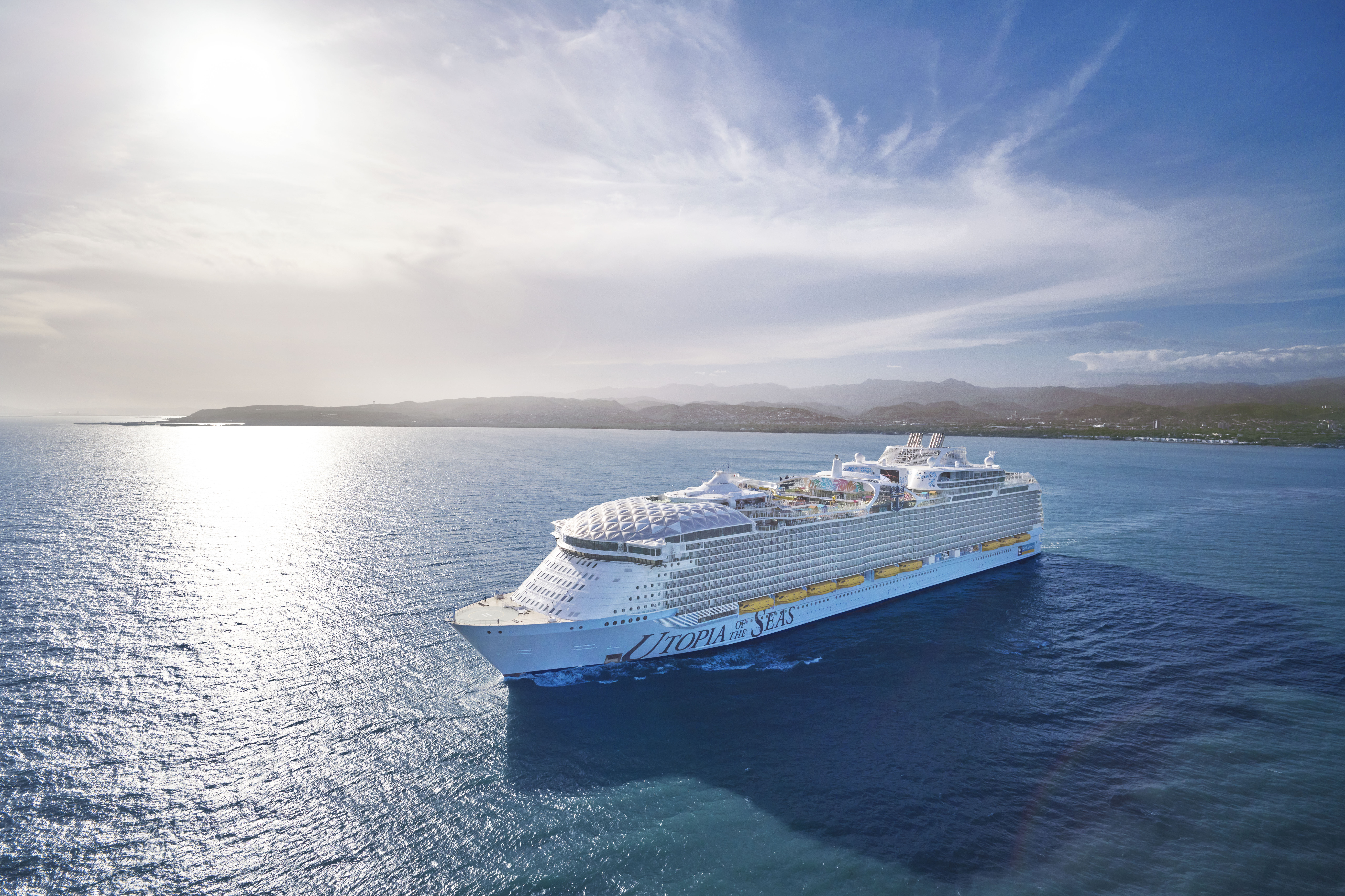 A stunning aerial photograph of a modern cruise ship departing from a tropical island port, showcasing the vessel's impressive white hull and multi-deck architecture against turquoise waters and volcanic island landscape under bright daylight.