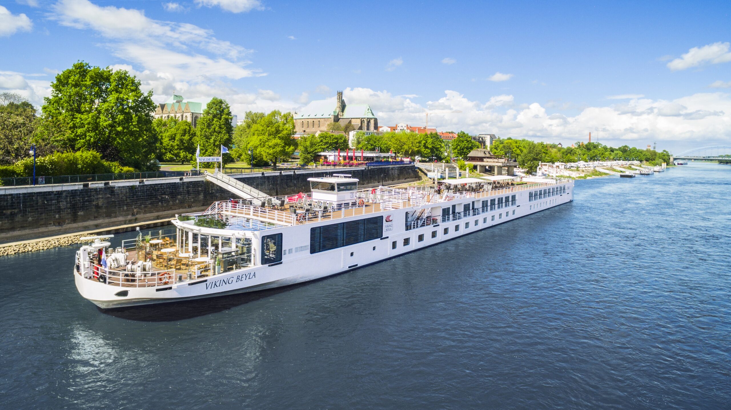 The Viking Beyla, a modern river cruise ship, navigates through Magdeburg, Germany, passing historic architecture and lush riverbanks along what appears to be the Elbe River. The elegant white vessel features multiple passenger decks and is docked near the city's medieval landmarks under a partly cloudy sky.