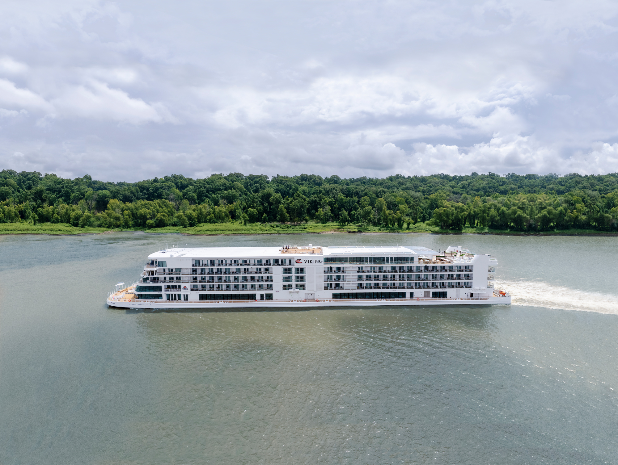 An aerial view of the Viking Mississippi, a modern river cruise ship, navigating the Mississippi River with dense green forests lining the riverbanks. The white multi-deck vessel is captured in profile on the calm, muddy-brown waters under a partly cloudy sky.