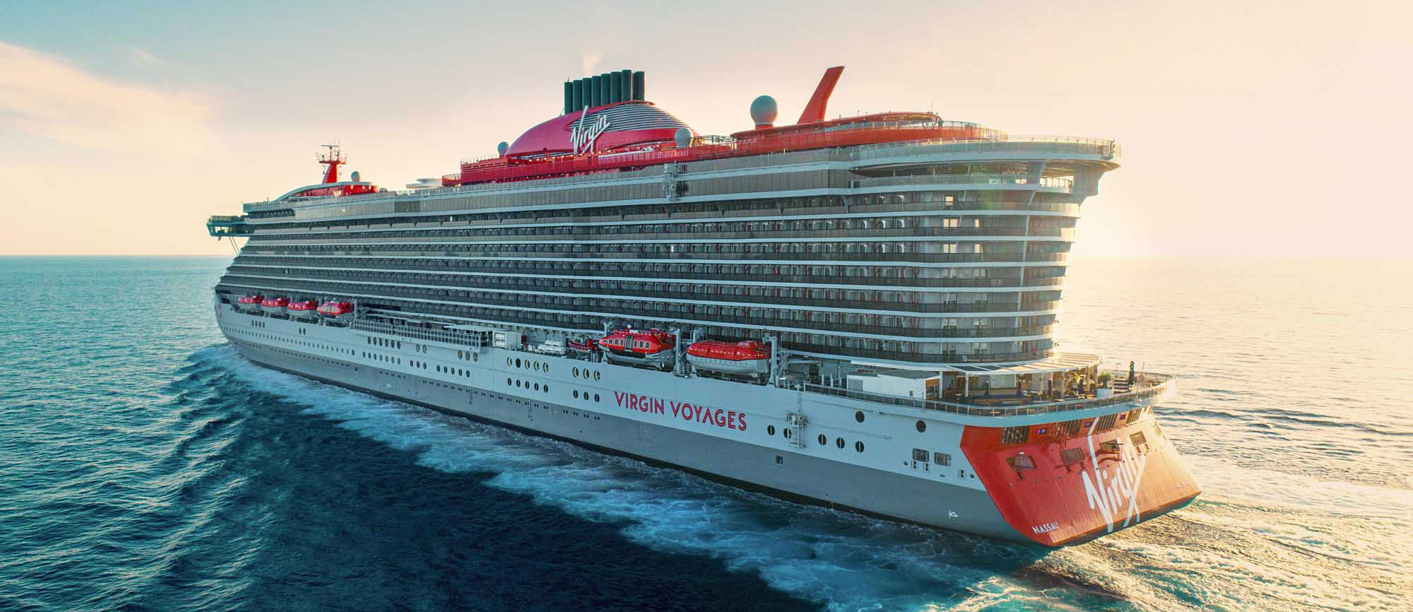 A modern cruise ship operated by Virgin Voyages is shown sailing in shallow coastal waters, displaying the distinctive red hull and white superstructure characteristic of the cruise line. The vessel features Virgin's signature red funnel with the 'V' logo and appears to be near a beach or shallow port area.