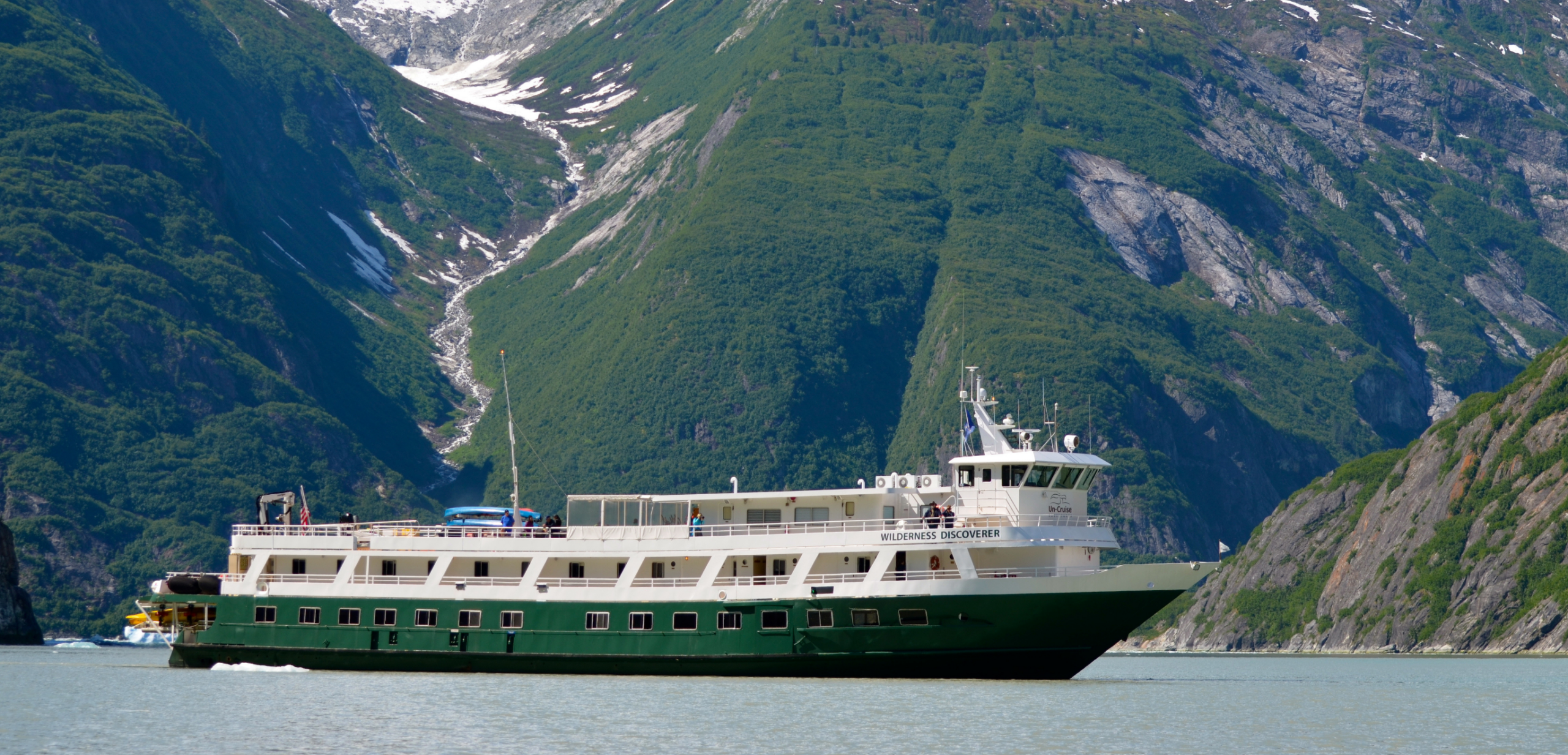 Wilderness Discoverer in Glacier Bay Alaska The small cruise ship Wilderness Discoverer navigates through a pristine Alaskan fjord with steep forested mountainsides, glaciers, and waterfalls visible in the background. The vessel, with its distinctive white upper decks and green hull, is anchored in calm, shallow waters typical of Southeast Alaska's Inside Passage.