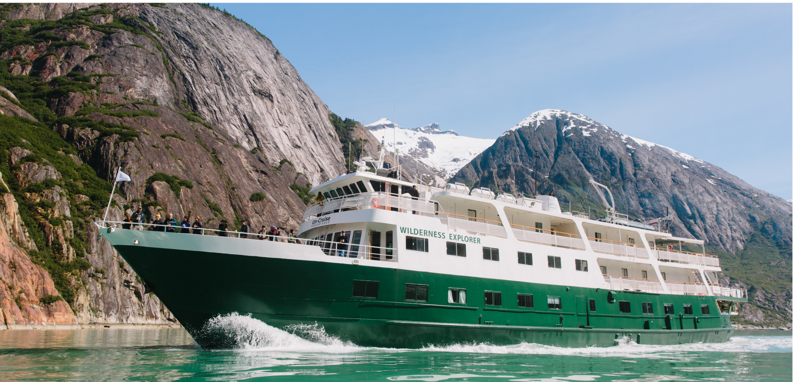 Wilderness Explorer Cruise Ship in Glacier Bay The Wilderness Explorer, a small expedition cruise ship with distinctive green and white hull, navigates through pristine glacial waters near towering snow-capped mountains and dramatic rocky cliffs. The vessel is positioned close to a rugged mountainside in what appears to be Alaska's Glacier Bay, showcasing the intimate exploration capabilities of small-ship cruising in remote polar regions.