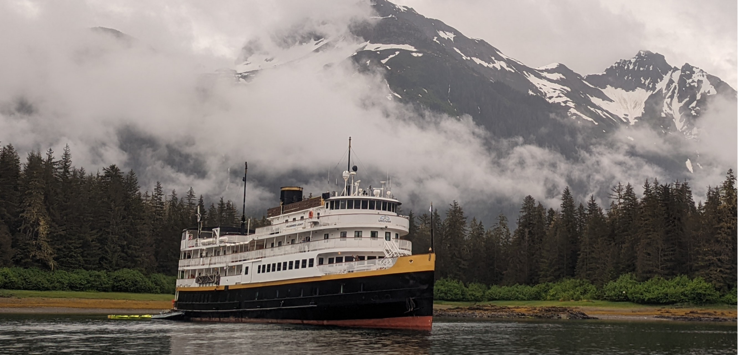 Wilderness Legacy Cruise Ship in Alaskan Waters A classic multi-deck cruise ship with white superstructure and black/red hull anchored in pristine Alaskan waters, surrounded by dense coniferous forests and snow-capped mountains shrouded in mist. The vessel features traditional maritime architecture with multiple decks, communication masts, and a distinctive funnel, embodying luxury small-ship cruising in remote wilderness destinations.