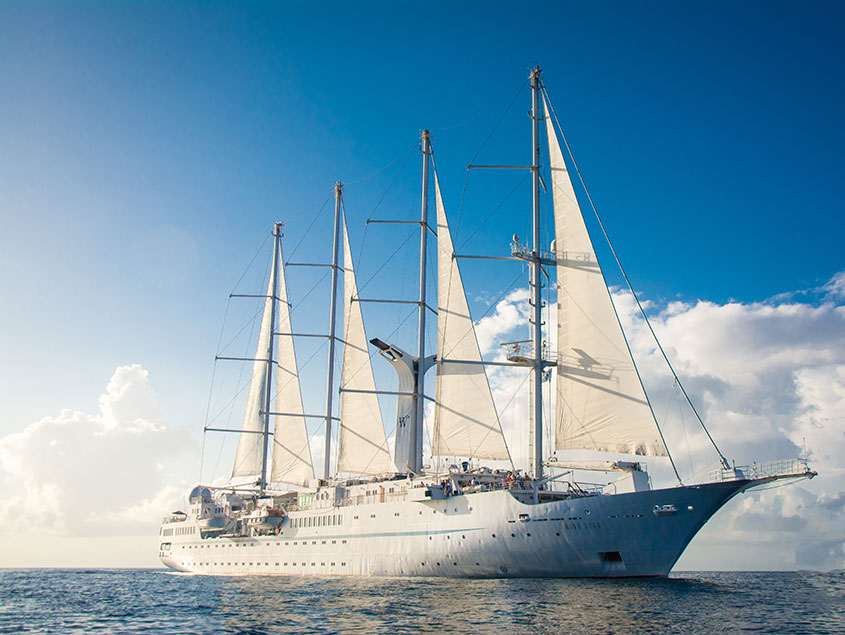 A luxurious small cruise ship with four tall masts and white sails fully deployed, sailing on calm blue ocean waters under clear skies. The vessel combines classic sailing ship aesthetics with modern cruise ship amenities, representing expedition and luxury travel.