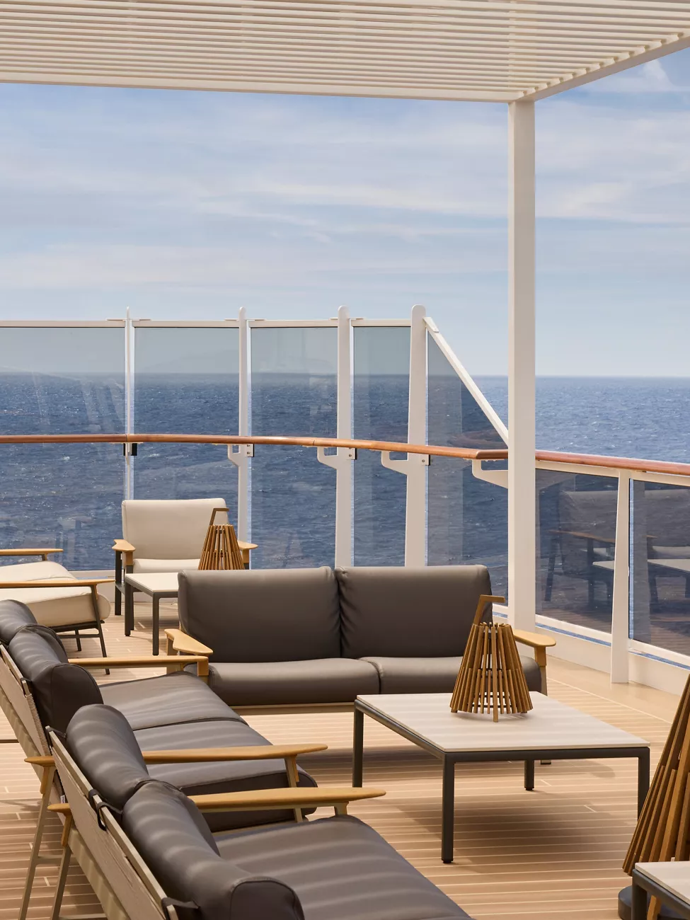 explora-sky-lounge-1 An elegant ship's sky lounge featuring contemporary furnishings with ocean views. The space includes comfortable seating arrangements with dark cushioned sofas and loungers, wooden accents, and floor-to-ceiling windows overlooking a calm sea beneath clear skies. A slatted ceiling provides partial shade while maintaining panoramic vistas.