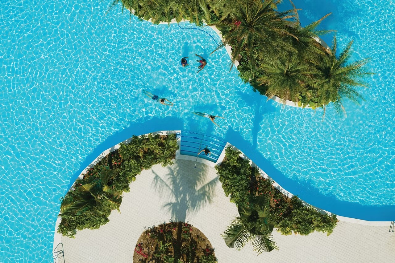 An overhead aerial view of a pristine turquoise lagoon pool deck featuring multiple swimmers, lush
