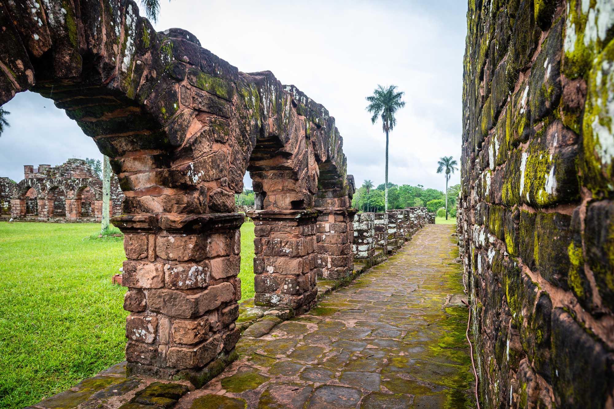 Ancient stone columns and archways covered in moss stand along a brick pathway at the ruins
