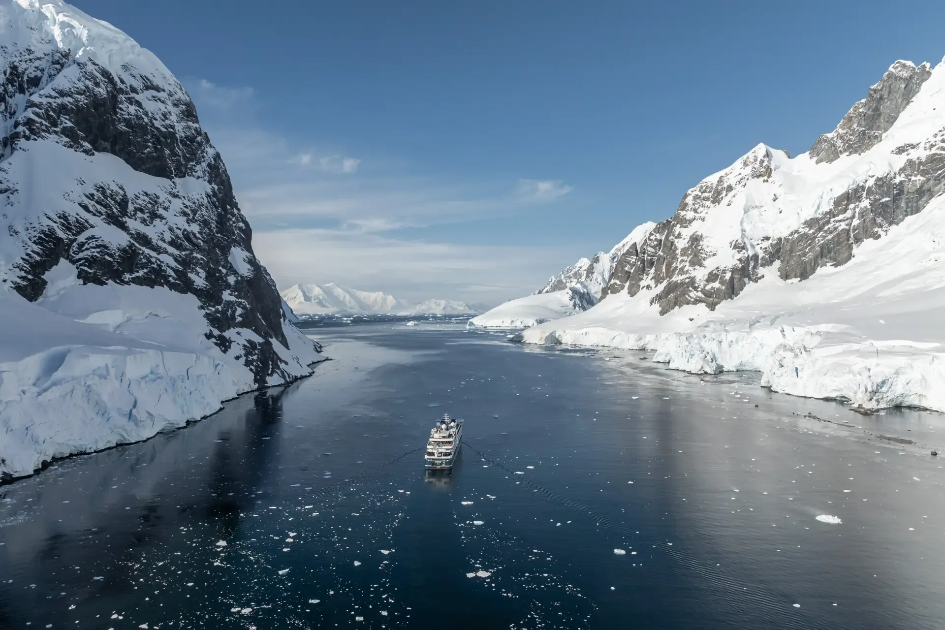 Expedition cruise ship navigating Antarctic channel between snow-covered mountain peaks and icebergs