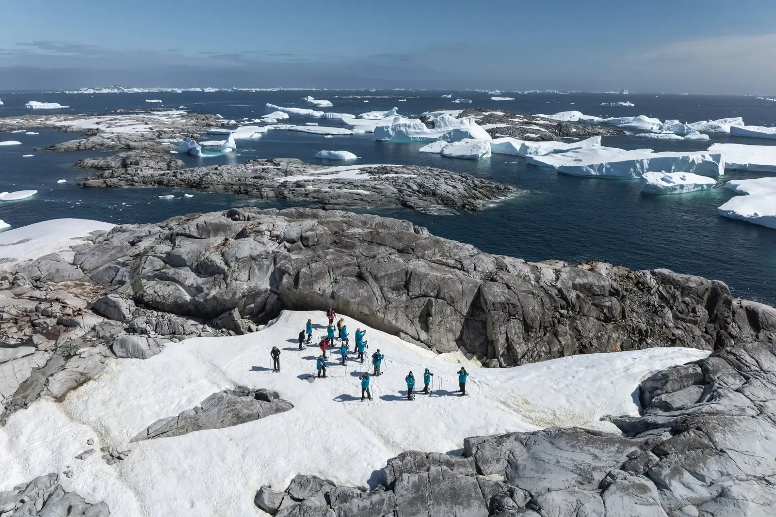 Expedition team in teal outfits on Greenland glacier surrounded by icebergs and rocky terrain