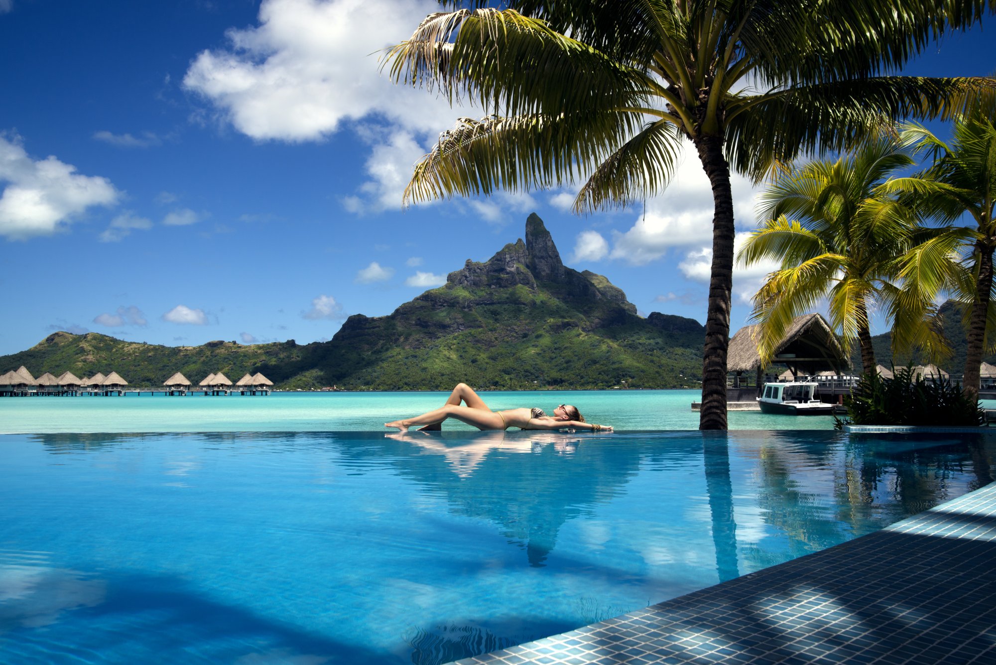 Woman relaxing at infinity pool edge with turquoise lagoon and volcanic mountain backdrop