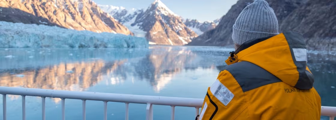 A bundled passenger in a bright yellow expedition jacket and gray beanie stands at the railing viewing a glacier at Alpefjord, East Greenland.