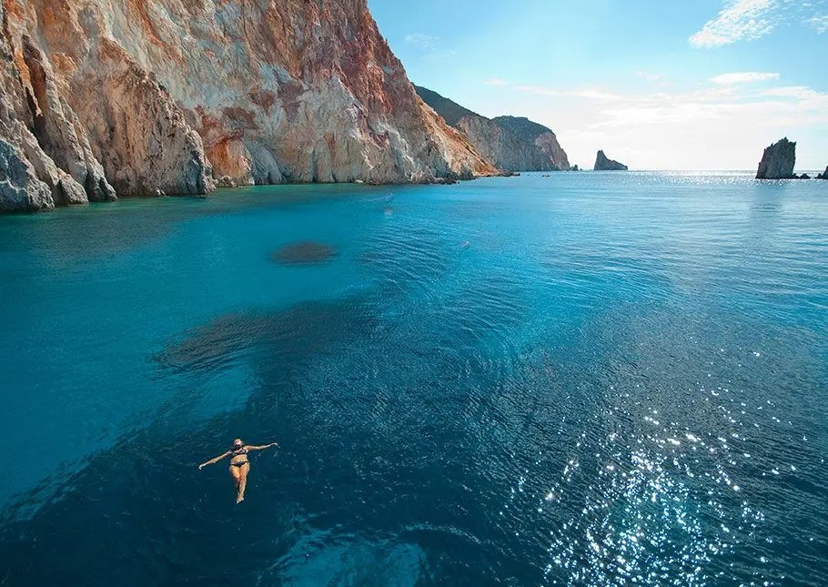 A swimmer floats in pristine turquoise Mediterranean waters beneath towering rust-colored Greek sea cliffs