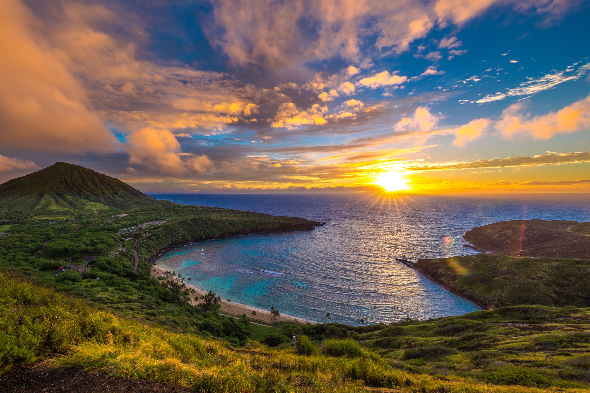 Sunset over turquoise Hawaiian cove with volcanic cliffs and palm-lined beach