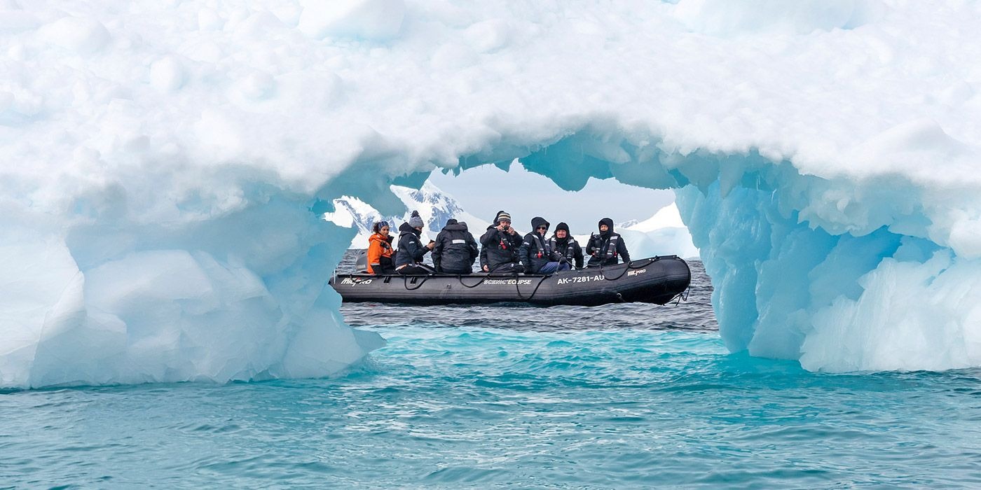 A group of expedition passengers navigate through a natural ice arch in a black Zodiac rigid