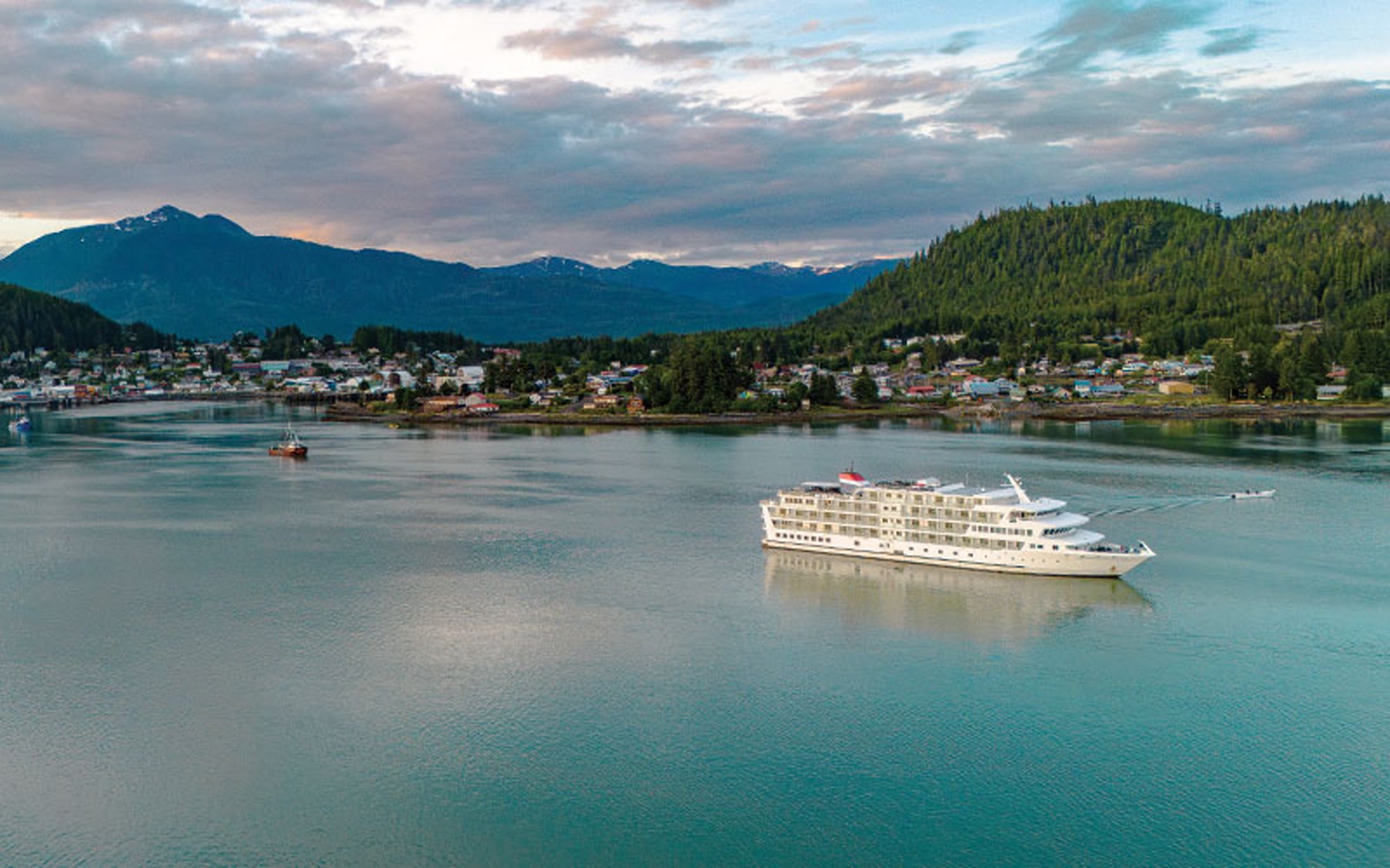 A large cruise ship is anchored in calm turquoise waters off a scenic Alaskan coastal community