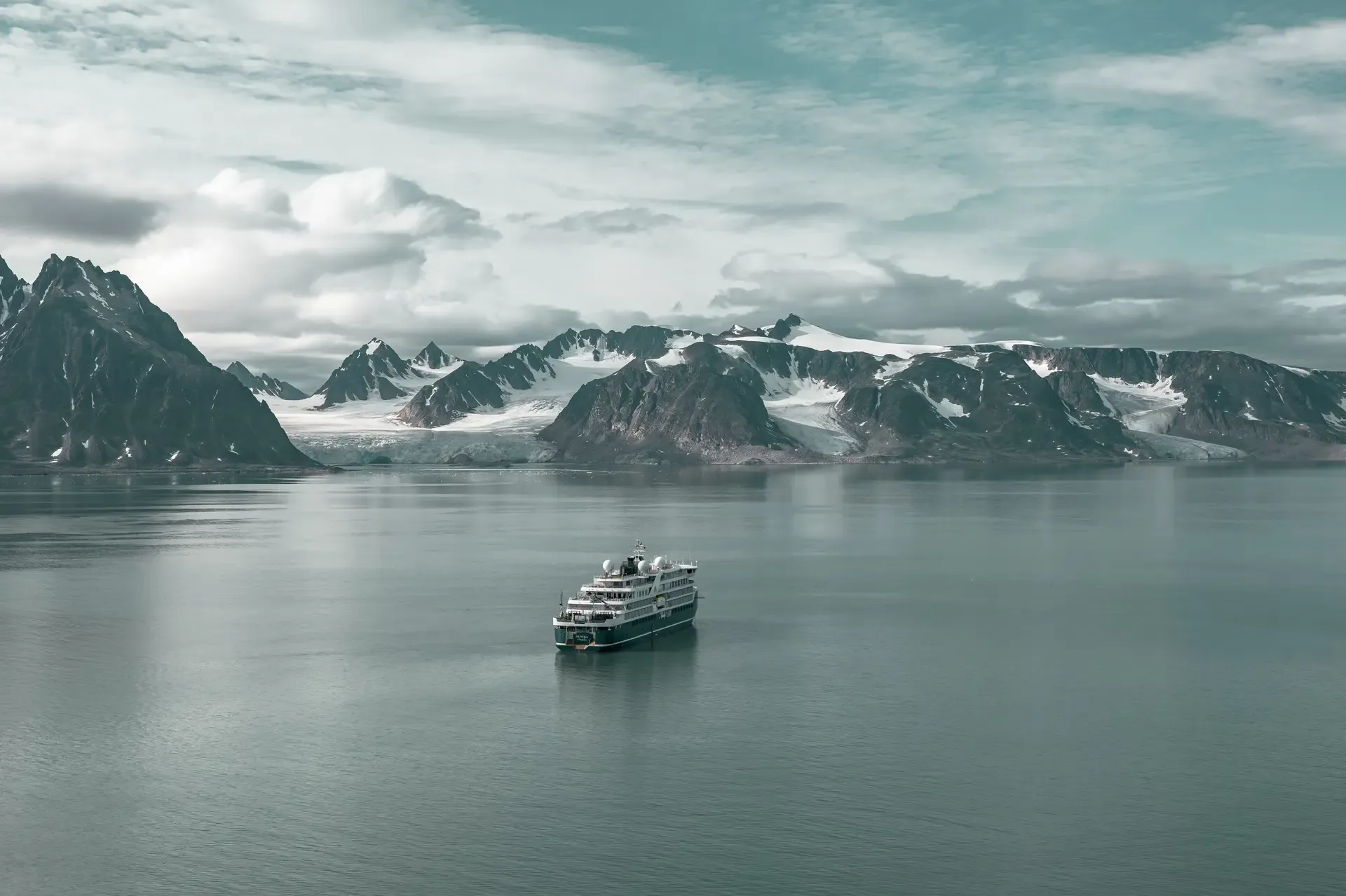A modern expedition cruise ship navigates calm Arctic waters surrounded by snow-capped mountains