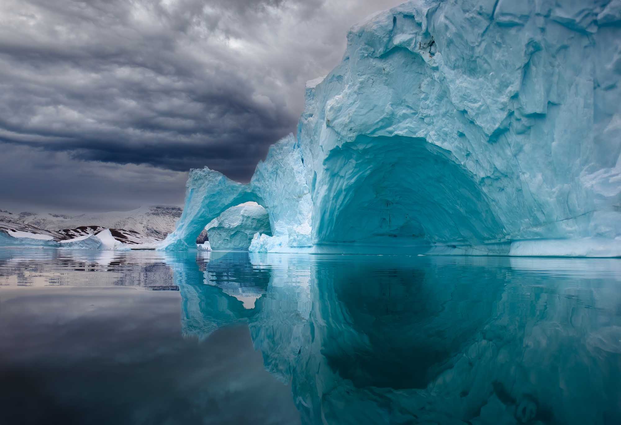 Iceberg Arch Reflected in East Greenland Waters A stunning natural ice arch formation in East Greenland, with turquoise glacial ice towering above