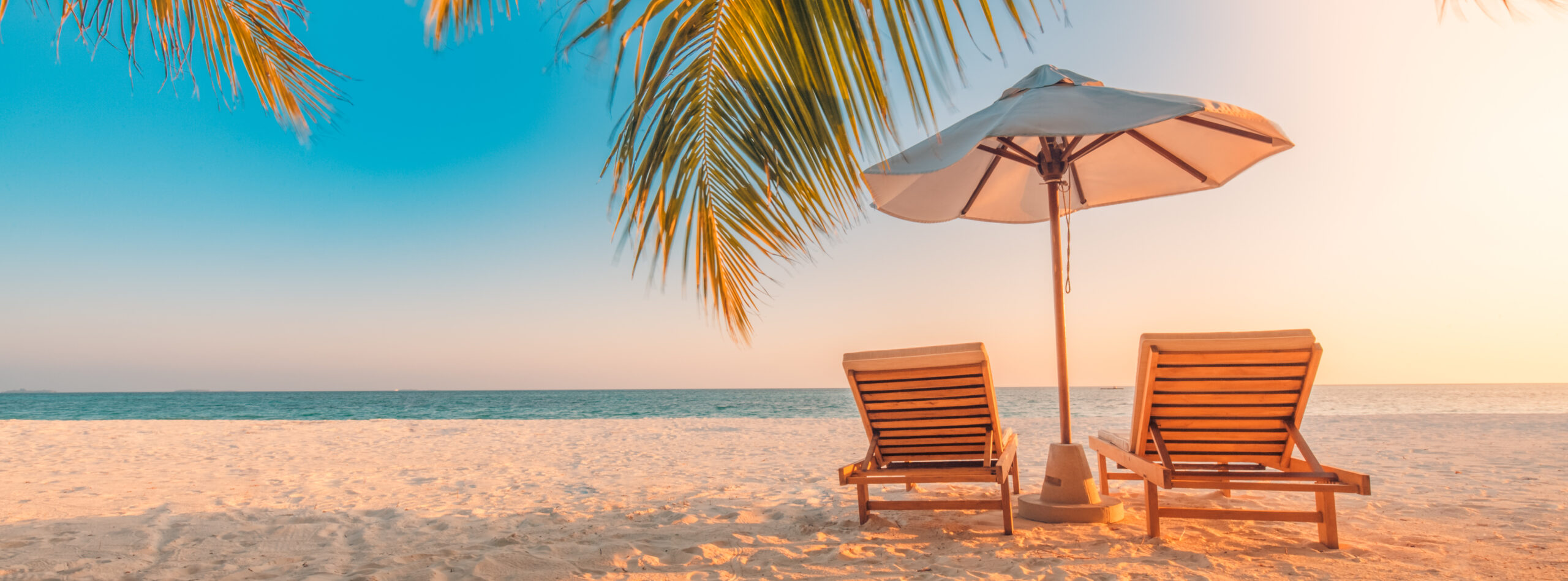 A serene tropical beach scene at golden hour featuring two wooden lounge chairs beneath a beige