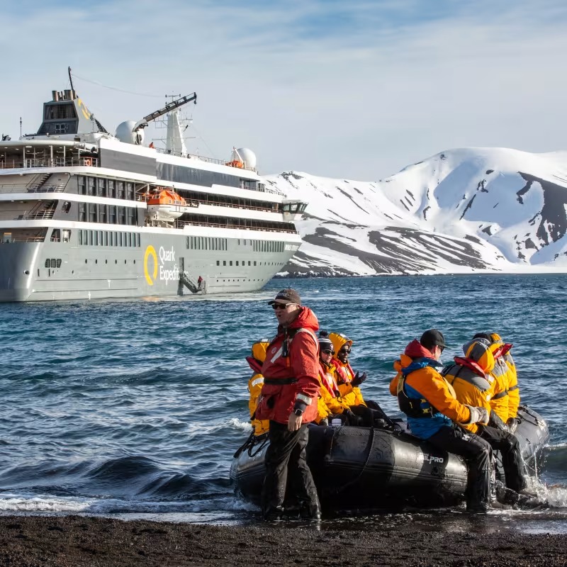 Passengers in bright yellow life jackets board a black inflatable Zodiac boat at the water's edge
